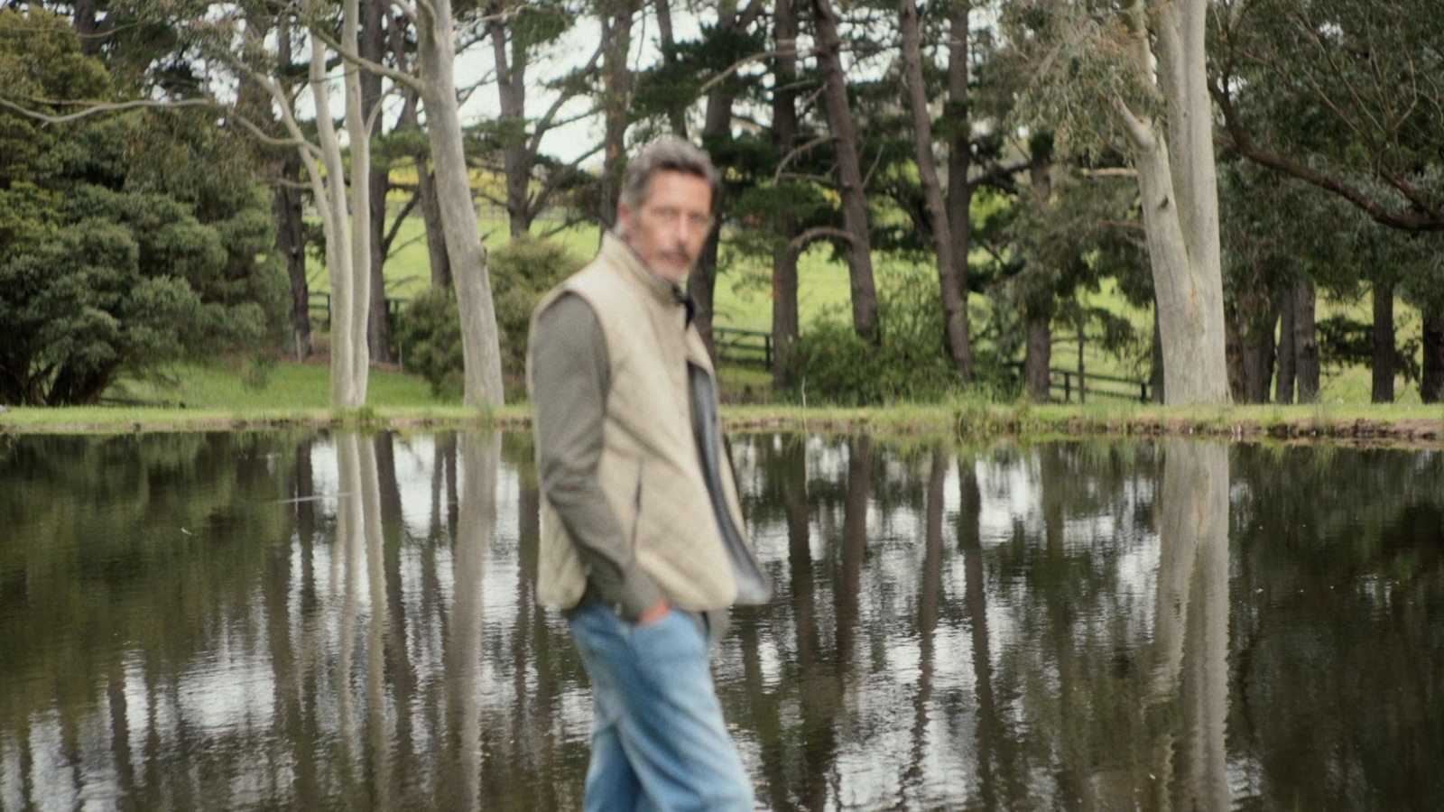 A man in a vest standing beside a still pond, trees reflected