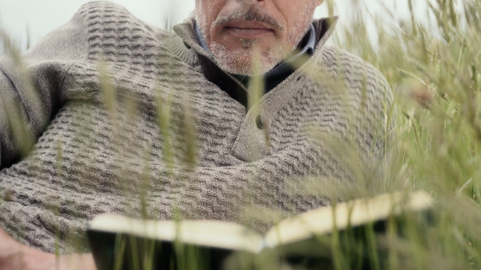 A man in a knit lying in long grass with a notebook